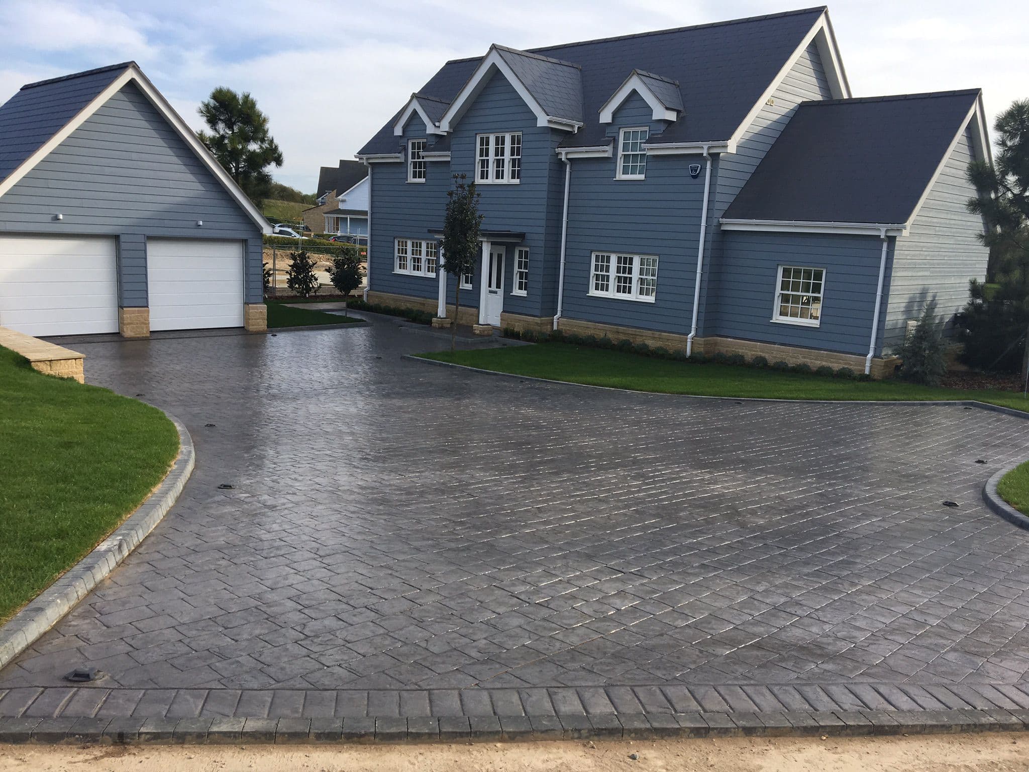 Large grey pattern imprinted concrete driveway laid in a cobble pattern with a darker stamped border, curving past a detached double garage to a weatherboarded house with lawn on either side