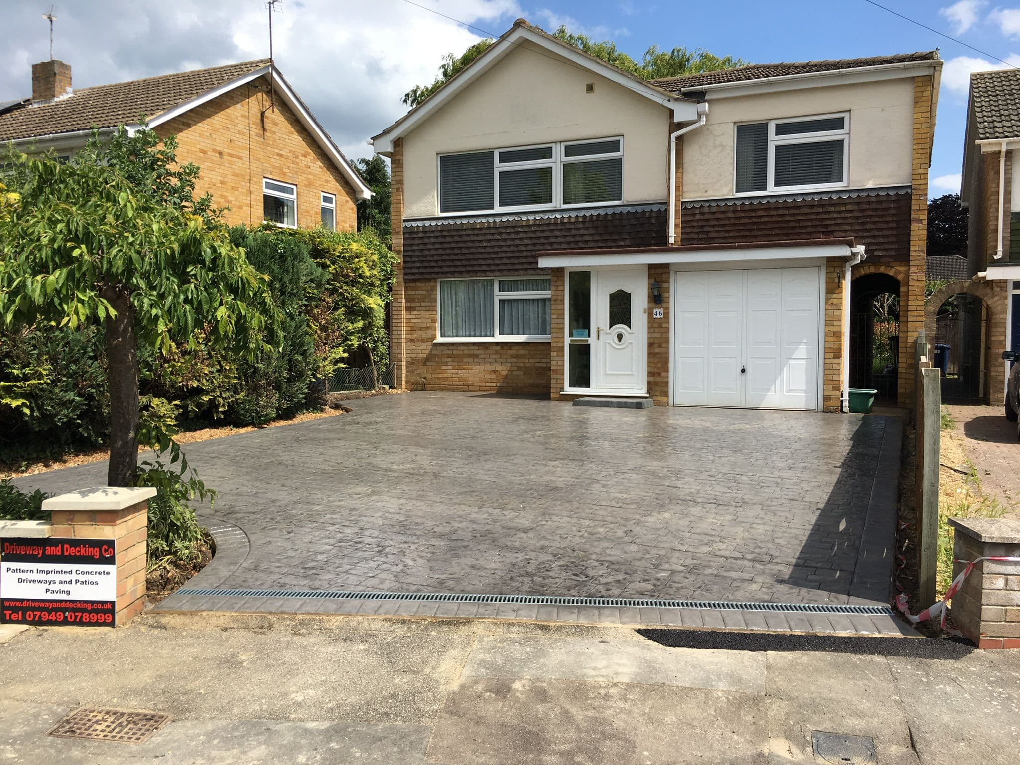 Mid grey ashlar pattern imprinted concrete driveway filling the frontage of a detached house, with a darker stamped cobble border and a metal channel drain along the pavement edge