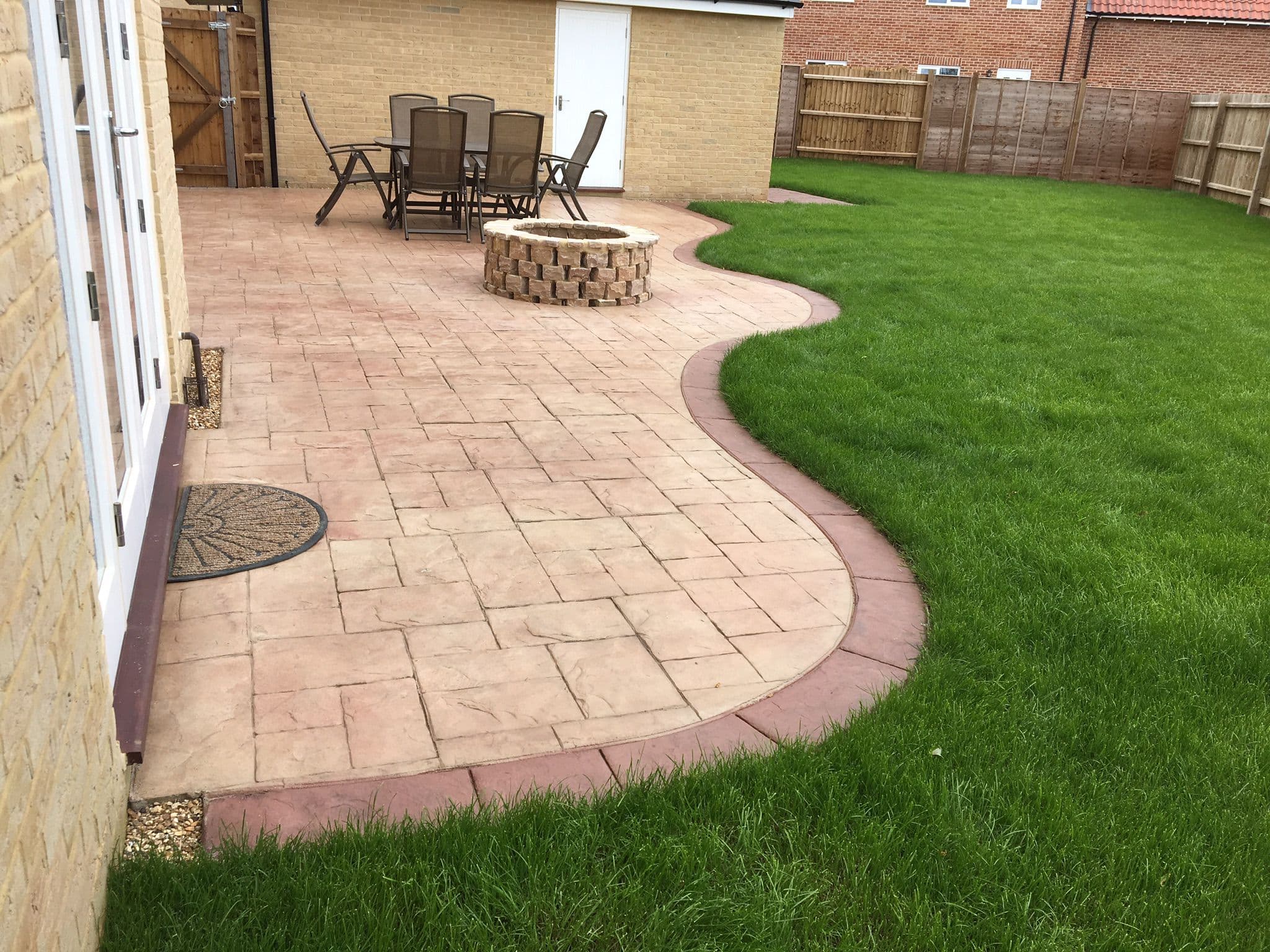 Buff ashlar pattern imprinted concrete patio with a curved terracotta red stamped border sweeping against a new lawn, with a circular brick fire pit and a garden dining set