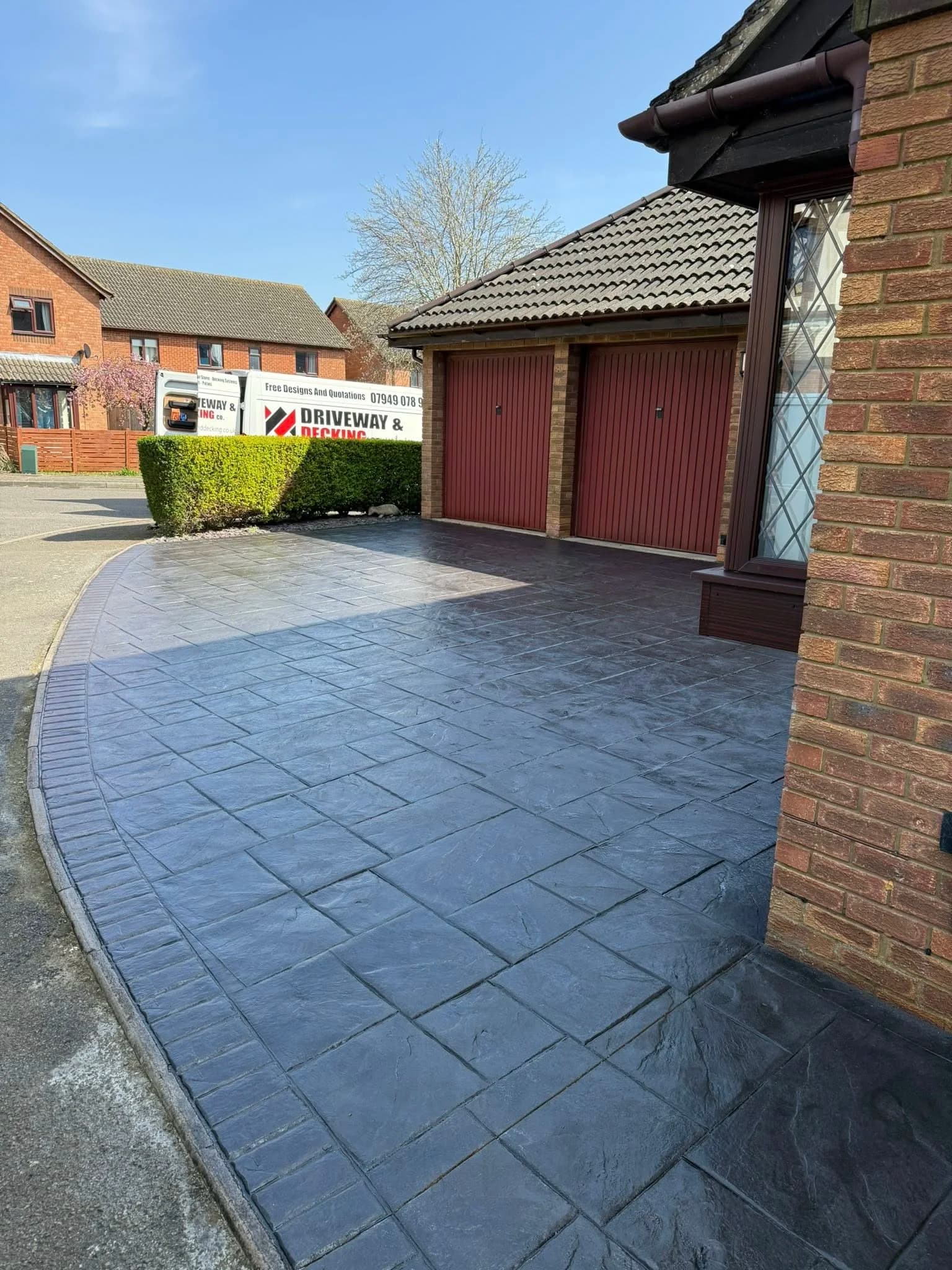 Dark charcoal ashlar pattern imprinted concrete driveway with a brick red stamped border, curving to a double garage with red up and over doors