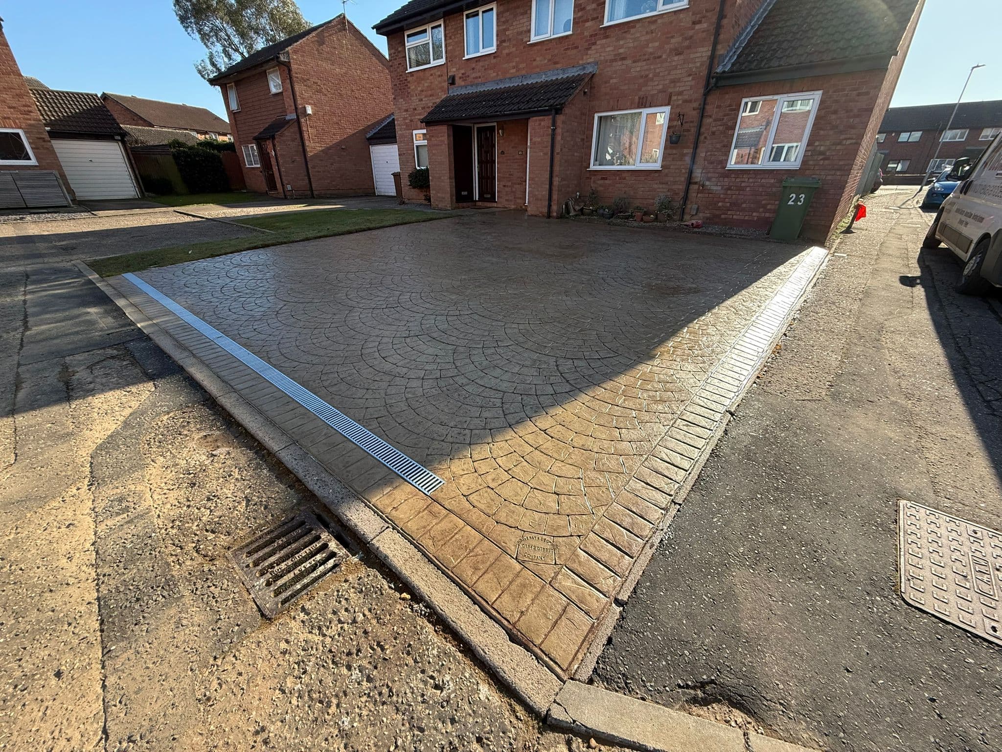 Buff coloured imprinted concrete driveway stamped in a circular European fan pattern, edged with a stamped brick soldier course and a metal channel drain, in front of a red brick house