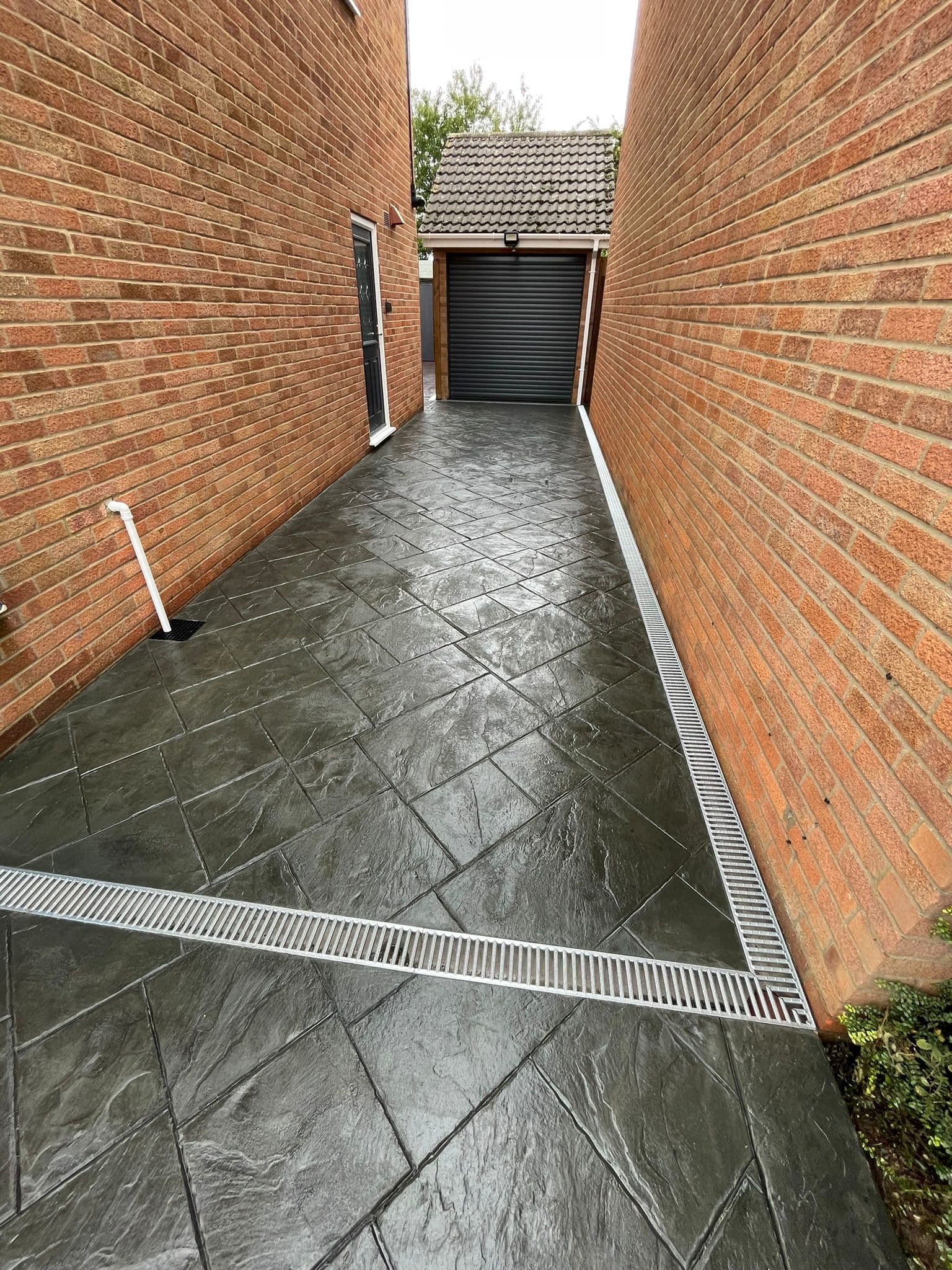 Dark charcoal imprinted concrete side path stamped in a riven stone pattern, running between two brick house walls to a grey roller garage door, with galvanised channel drains along the edge