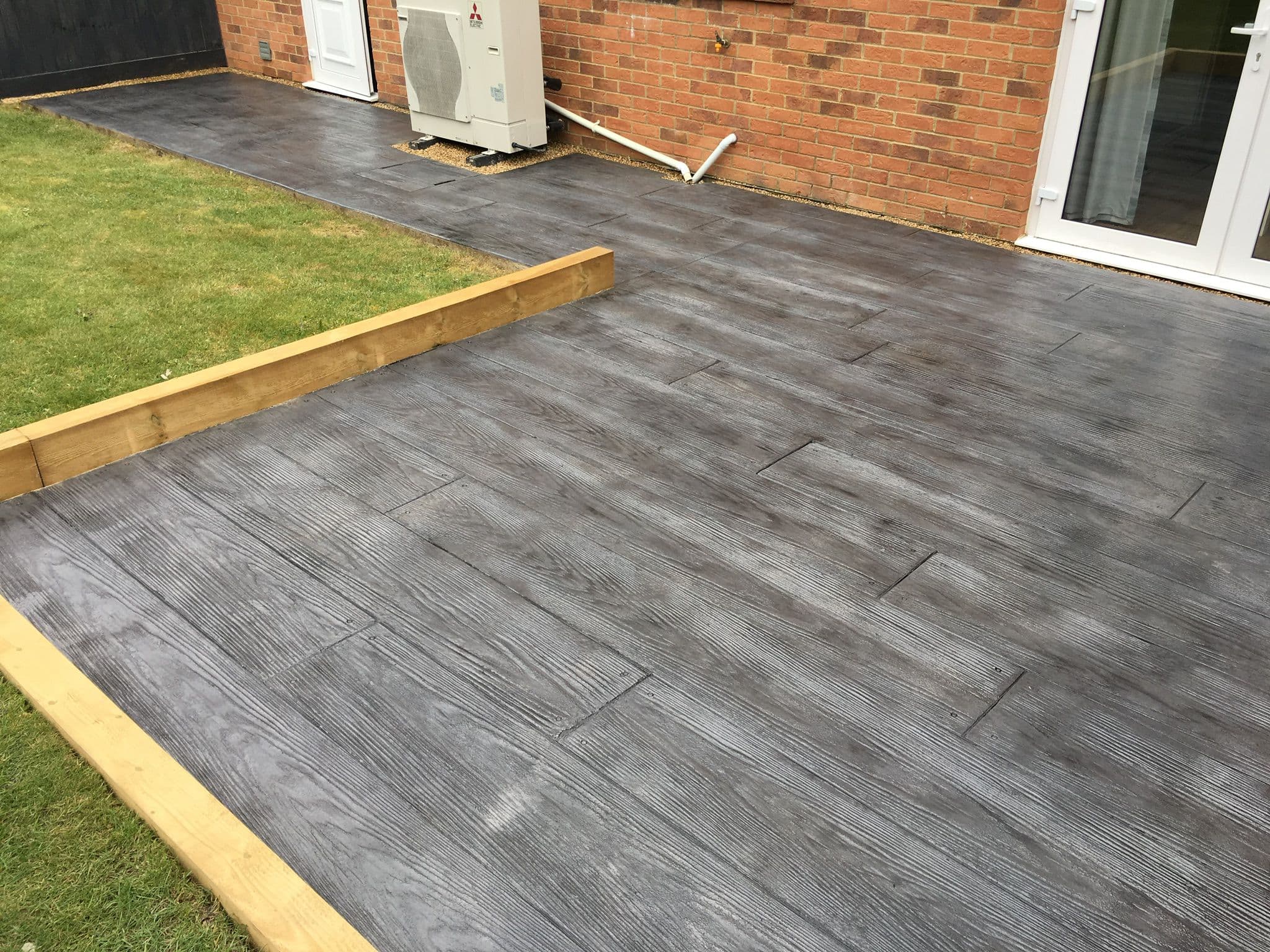 Grey imprinted concrete patio stamped to look like wide timber boards, with visible grain and nail head detail, edged with a timber sleeper against a lawn